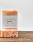 Two bars of artisan soap labeled 'renew' with bergamot and orange scent. The top bar has a smooth surface, while the bottom has a swirled design. They are placed on a wooden surface with a light background.