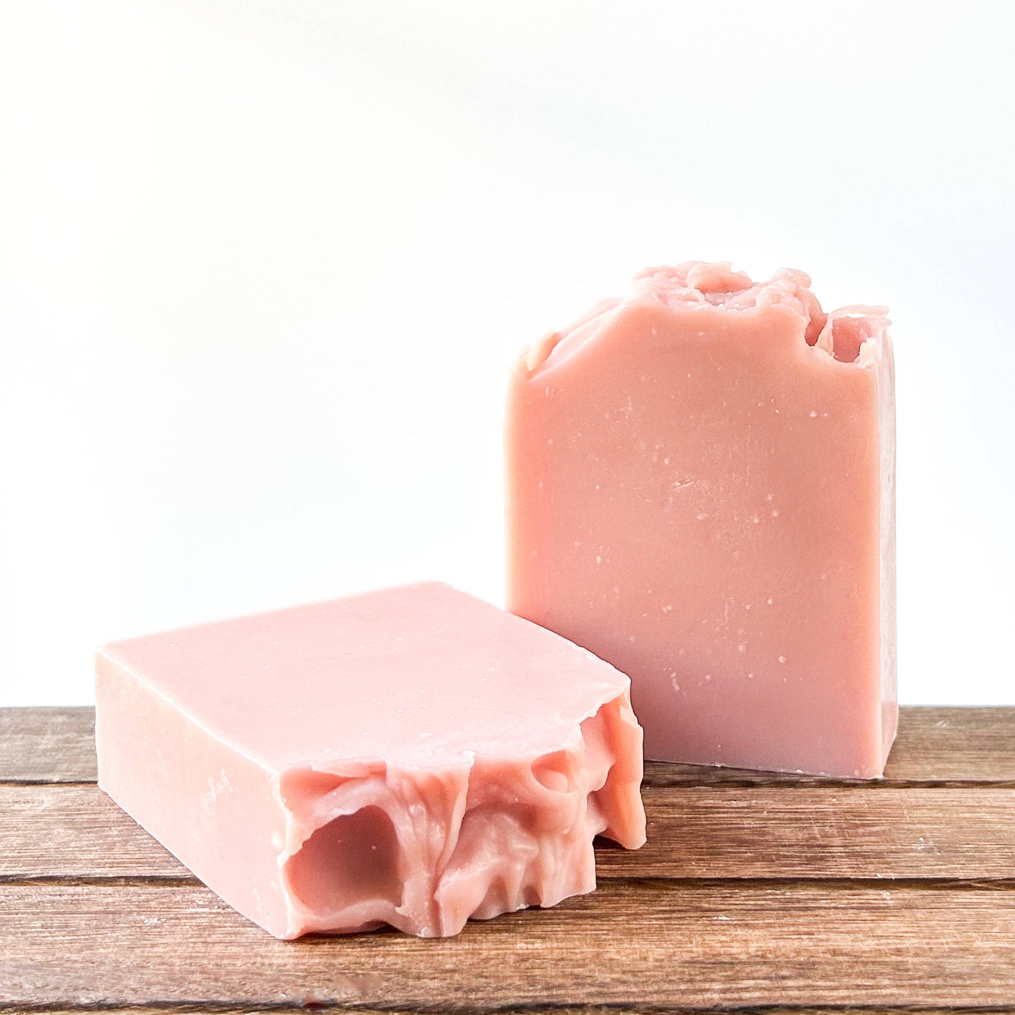 Two pieces of pink soap displayed on a wooden surface, with sculpted tops. The background is bright and minimalist.