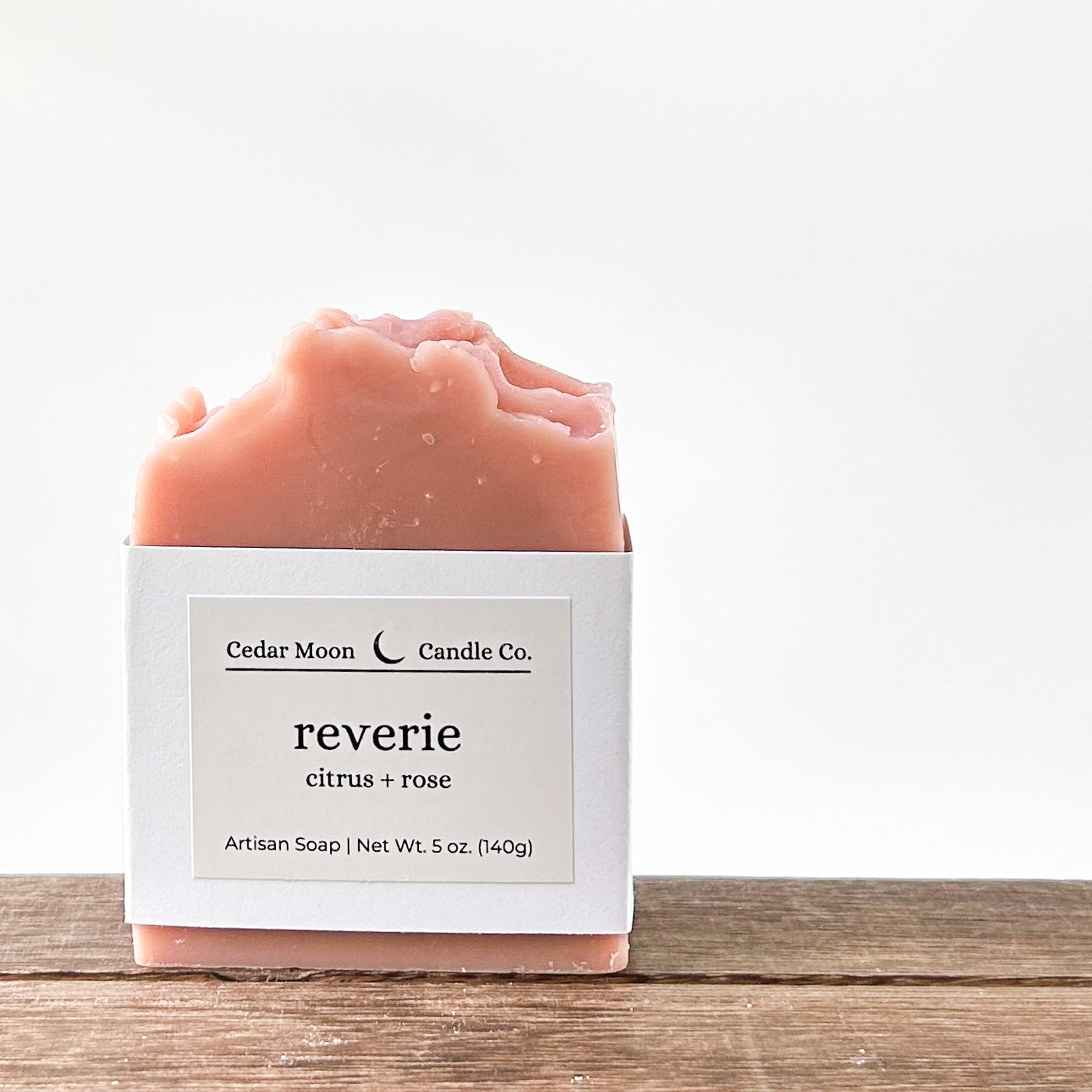A pink bar of artisan soap, labeled 'reverie citrus rose'. The soap is resting on a wooden surface with a minimalist background.