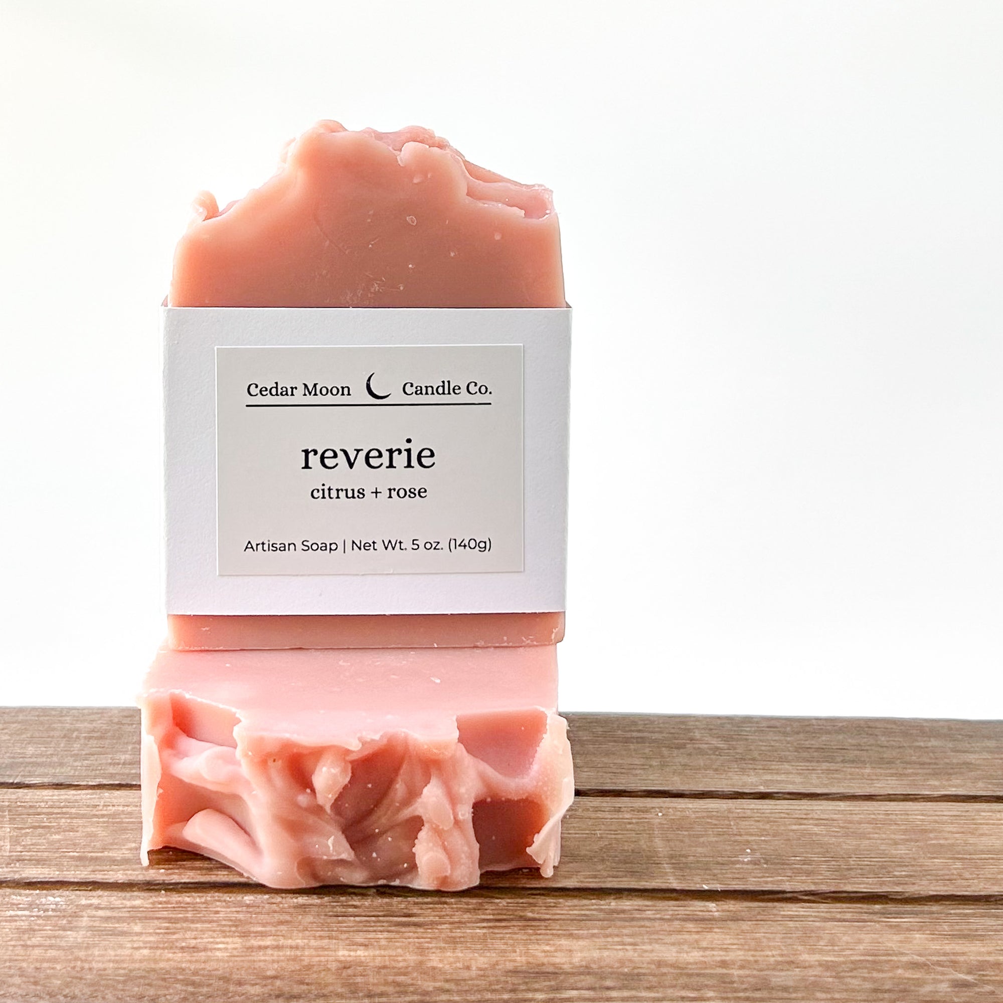 Artisan soap from Cedar Moon Candle Co. in a pink hue, labeled 'reverie citrus  rose'. The soap is on a wooden surface with a glossy finish, showcasing its texture.