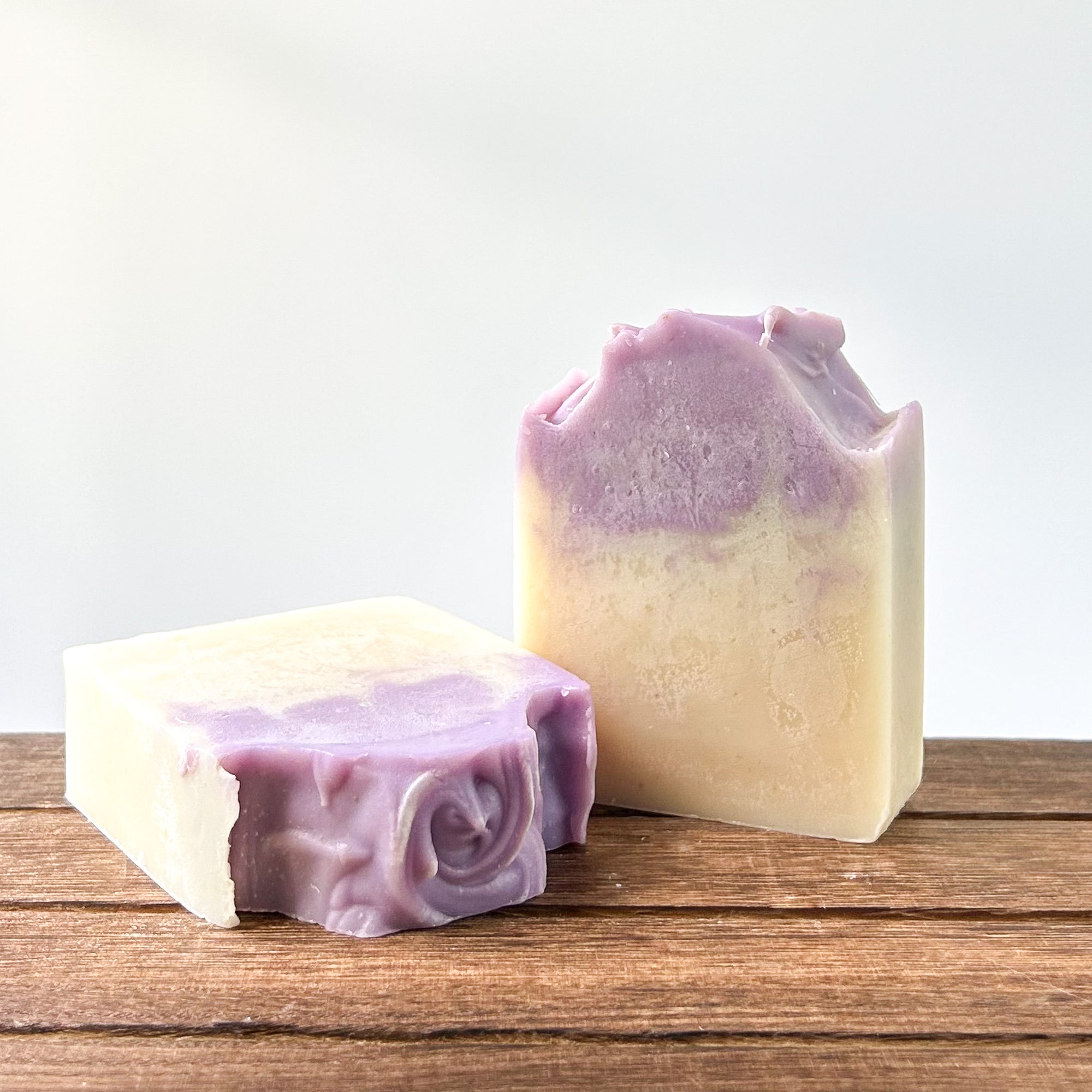 Two handcrafted soap bars in purple and cream colors, one with a decorative swirl design. They are placed on a wooden surface with a soft blurred background.