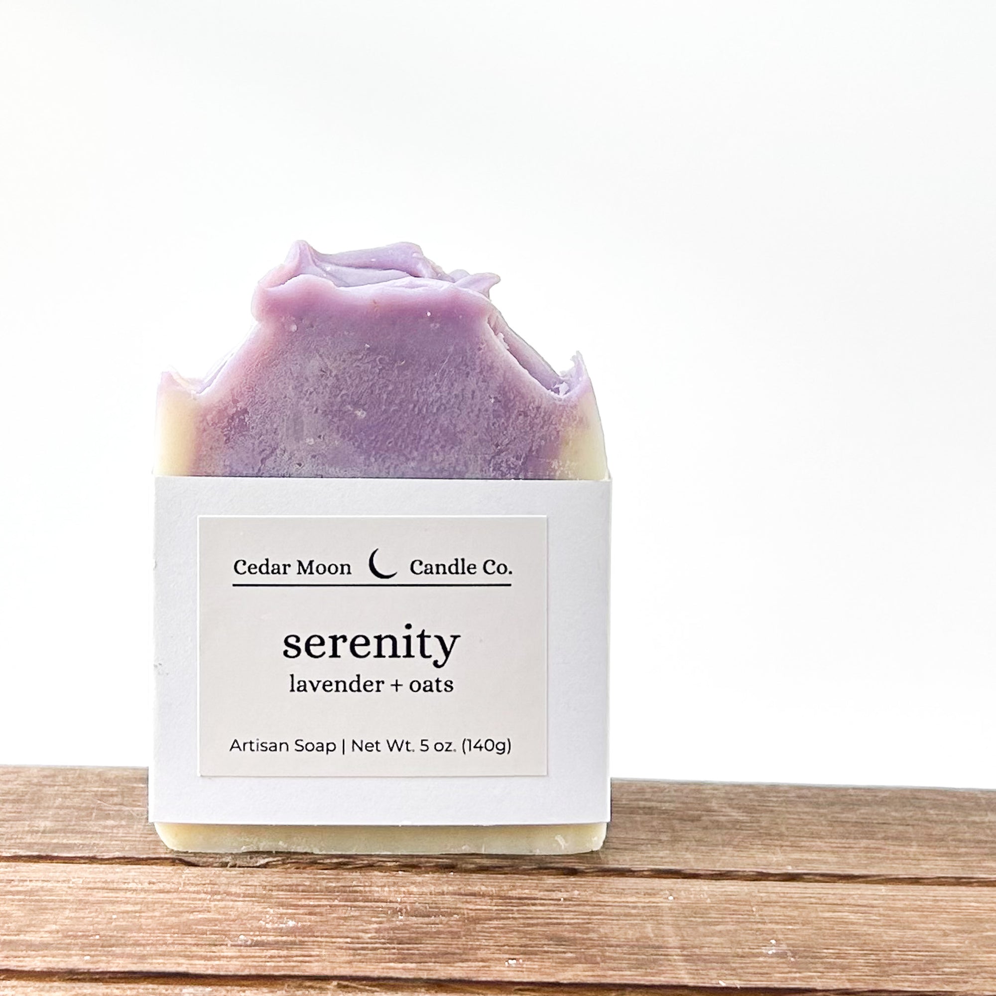 A bar of 'serenity' artisan soap featuring lavender and oats. The soap is a creamy white and purple swirl pattern, wrapped in a white paper band placed on a wooden surface against a light background.