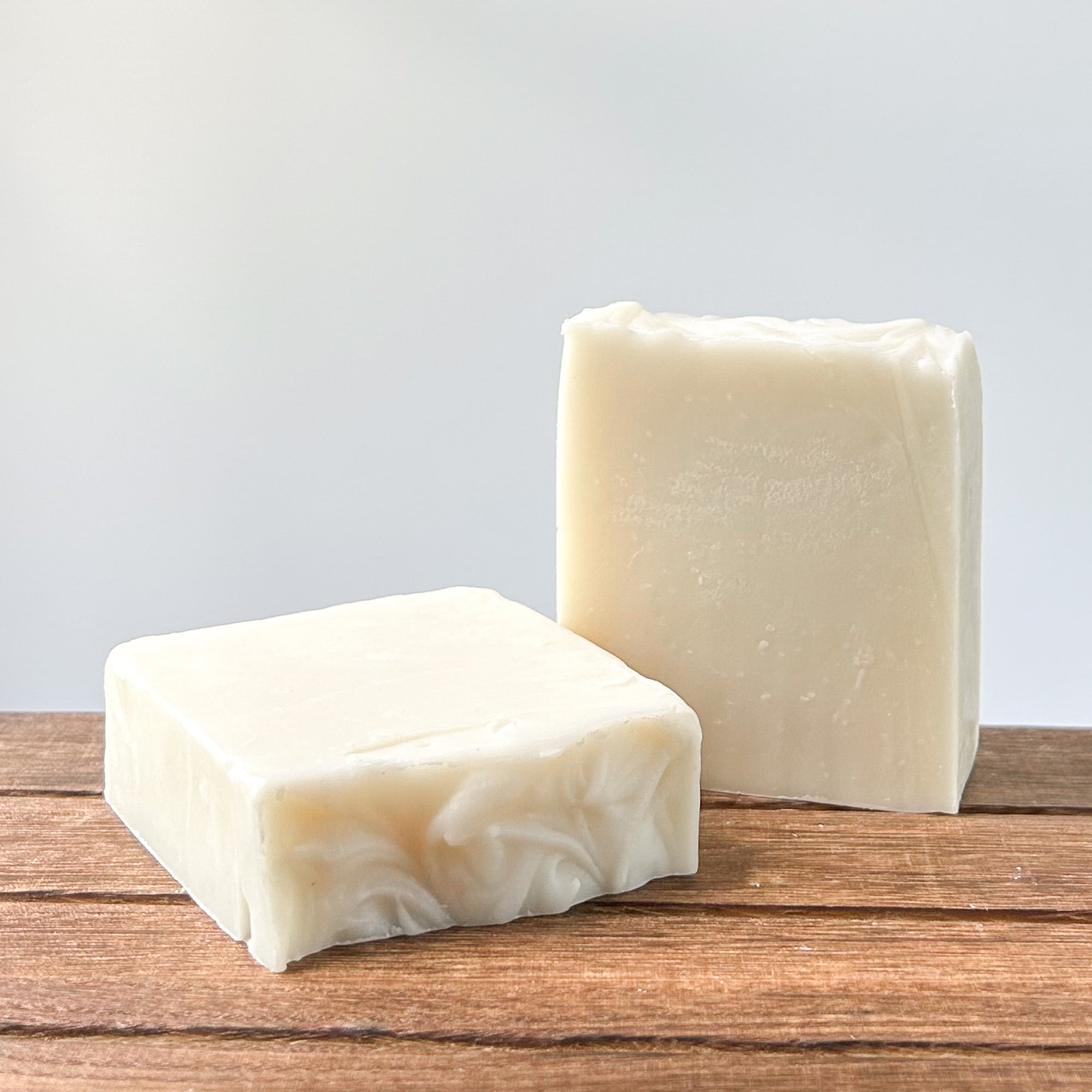 Two bars of handmade soap in white and light cream colors resting on a wooden surface, with a soft, neutral background.