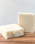 Two bars of handmade soap in white and light cream colors resting on a wooden surface, with a soft, neutral background.