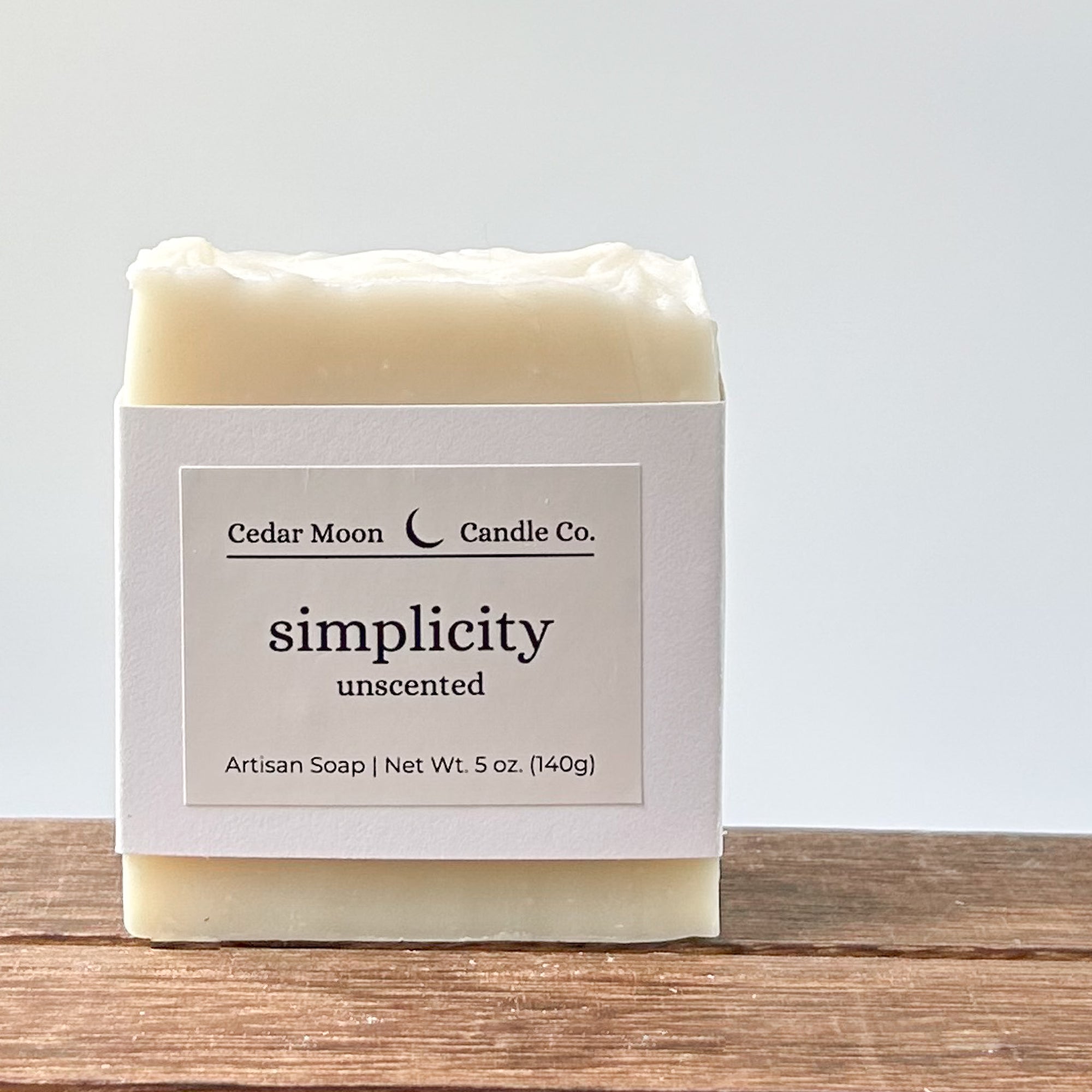 A bar of 'simplicity' unscented artisan soap. The soap is a creamy white and wrapped in a white paper band placed on a wooden surface against a light background.