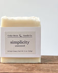 A bar of 'simplicity' unscented artisan soap. The soap is a creamy white and wrapped in a white paper band placed on a wooden surface against a light background.
