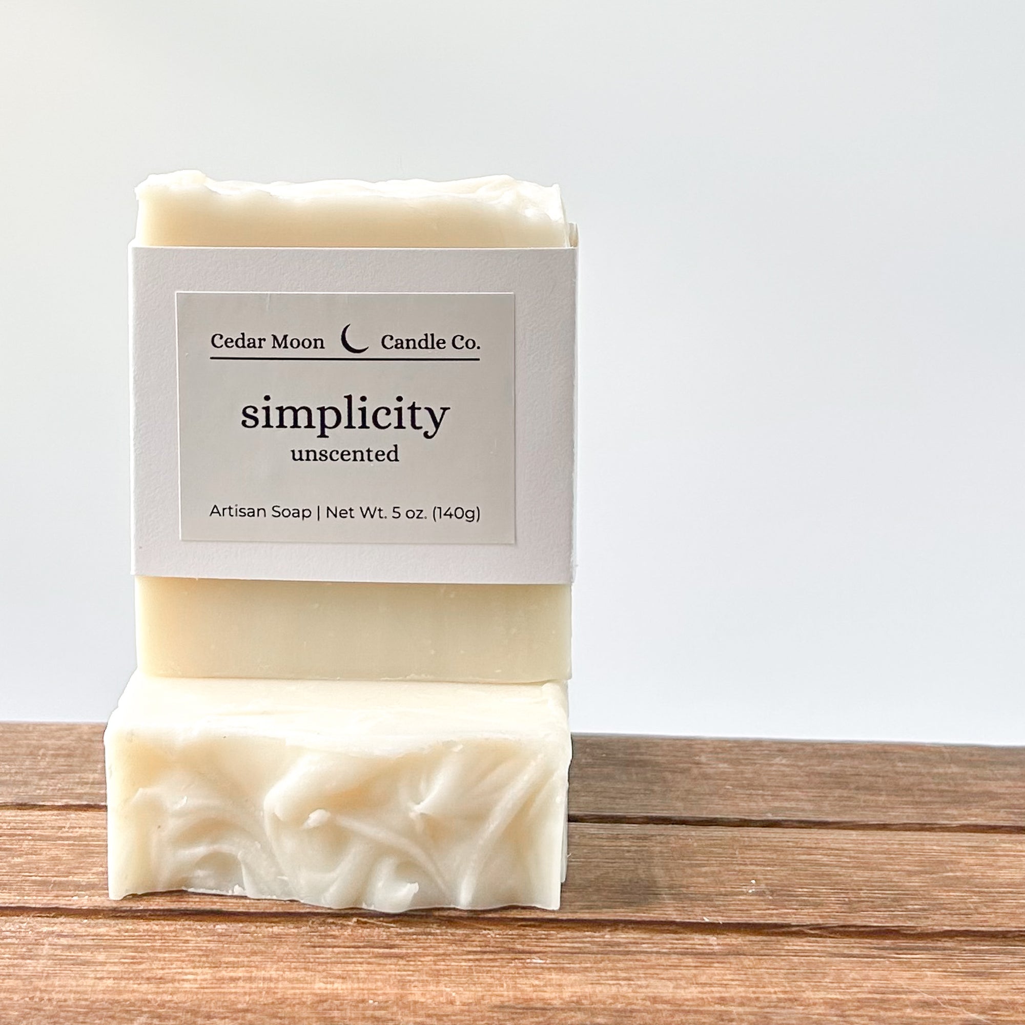 A close-up of an unscented artisan soap labeled 'simplicity' by Cedar Moon Candle Co. The soap is white with a textured surface, placed on a wooden surface with a light background.