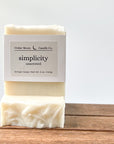 A close-up of an unscented artisan soap labeled 'simplicity' by Cedar Moon Candle Co. The soap is white with a textured surface, placed on a wooden surface with a light background.