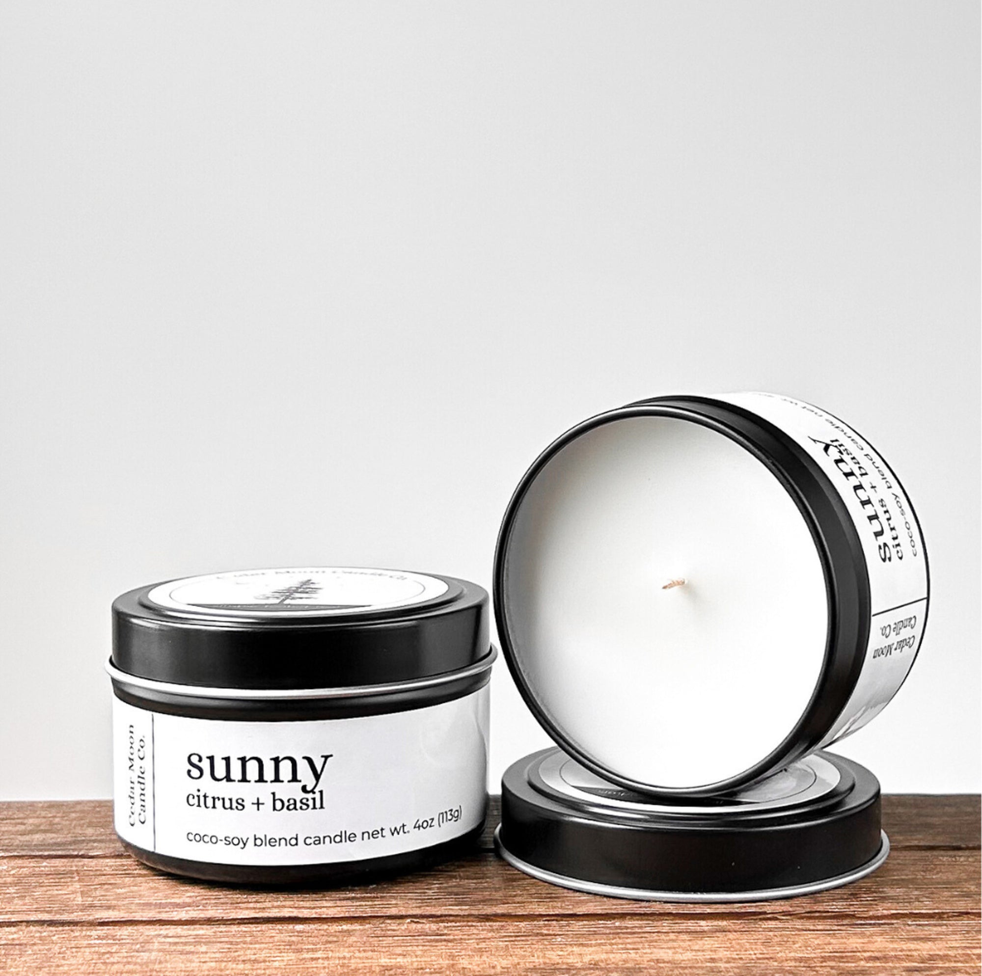 Two black tins of 'sunny citrus and basil' coco-soy blend candle, one lid off, showcasing the white wax and wick. Placed on a wooden surface with a neutral background.