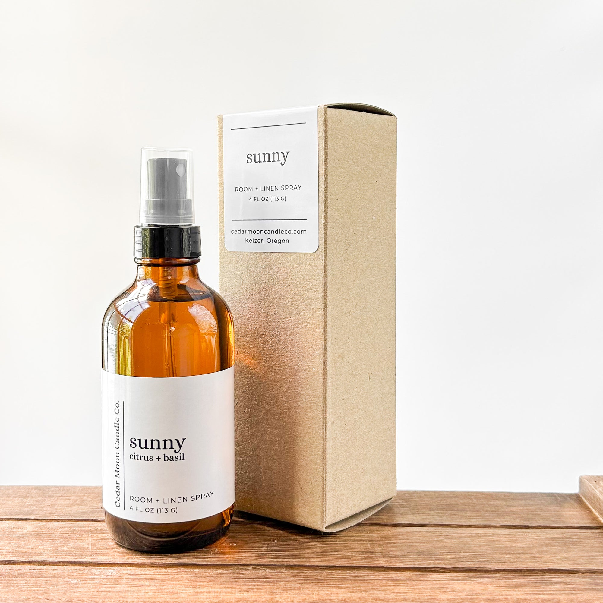 A bottle of room and linen spray labeled 'sunny citrus and basil' next to a simple cardboard box, resting on a wooden surface with a neutral background.