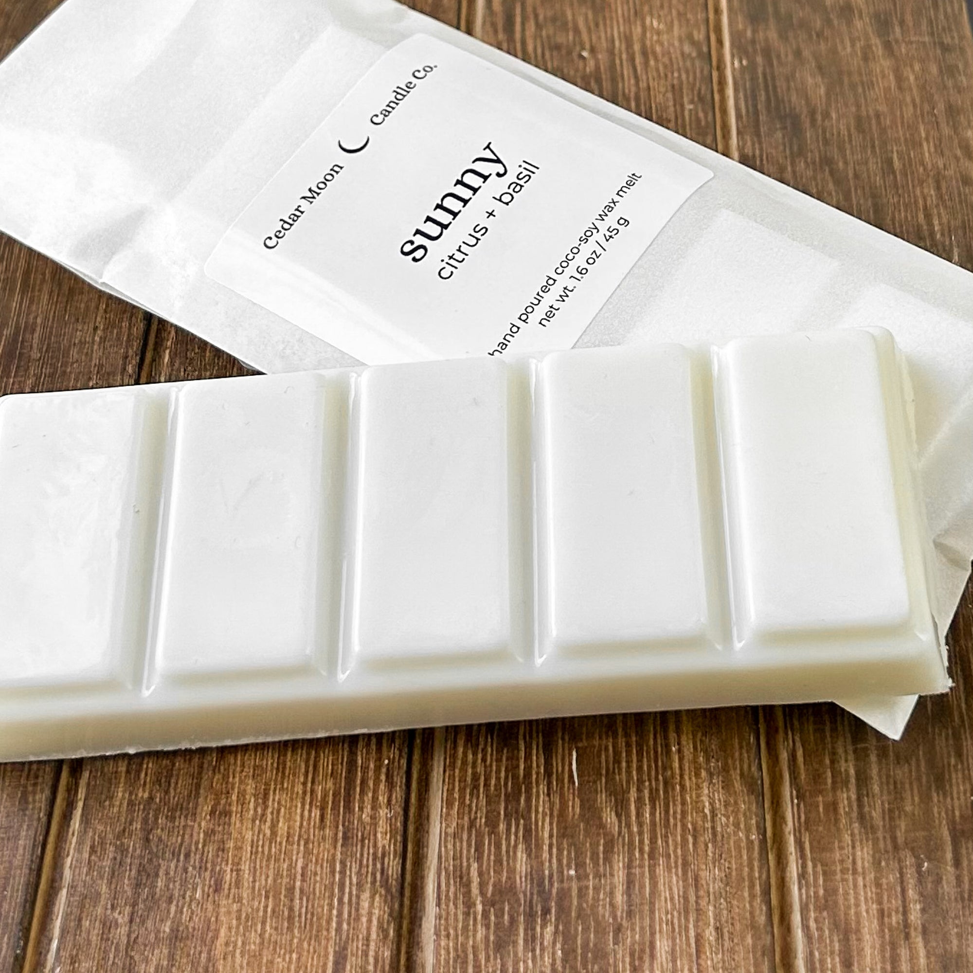 A white wax melt bar labeled 'sunny' with scents of citrus and basil, resting on a textured wooden surface. The package highlights the product's natural ingredients.