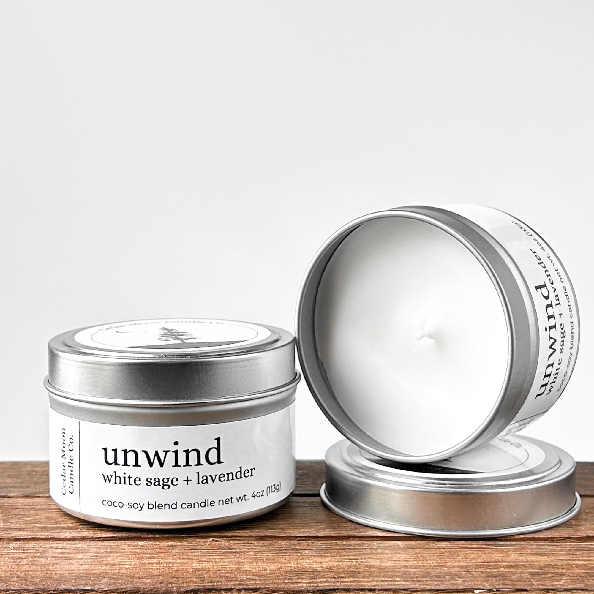 Two silver tins of 'unwind white sage and lavender' coco-soy blend candle, one lid off, showcasing the white wax and wick. Placed on a wooden surface with a neutral background.