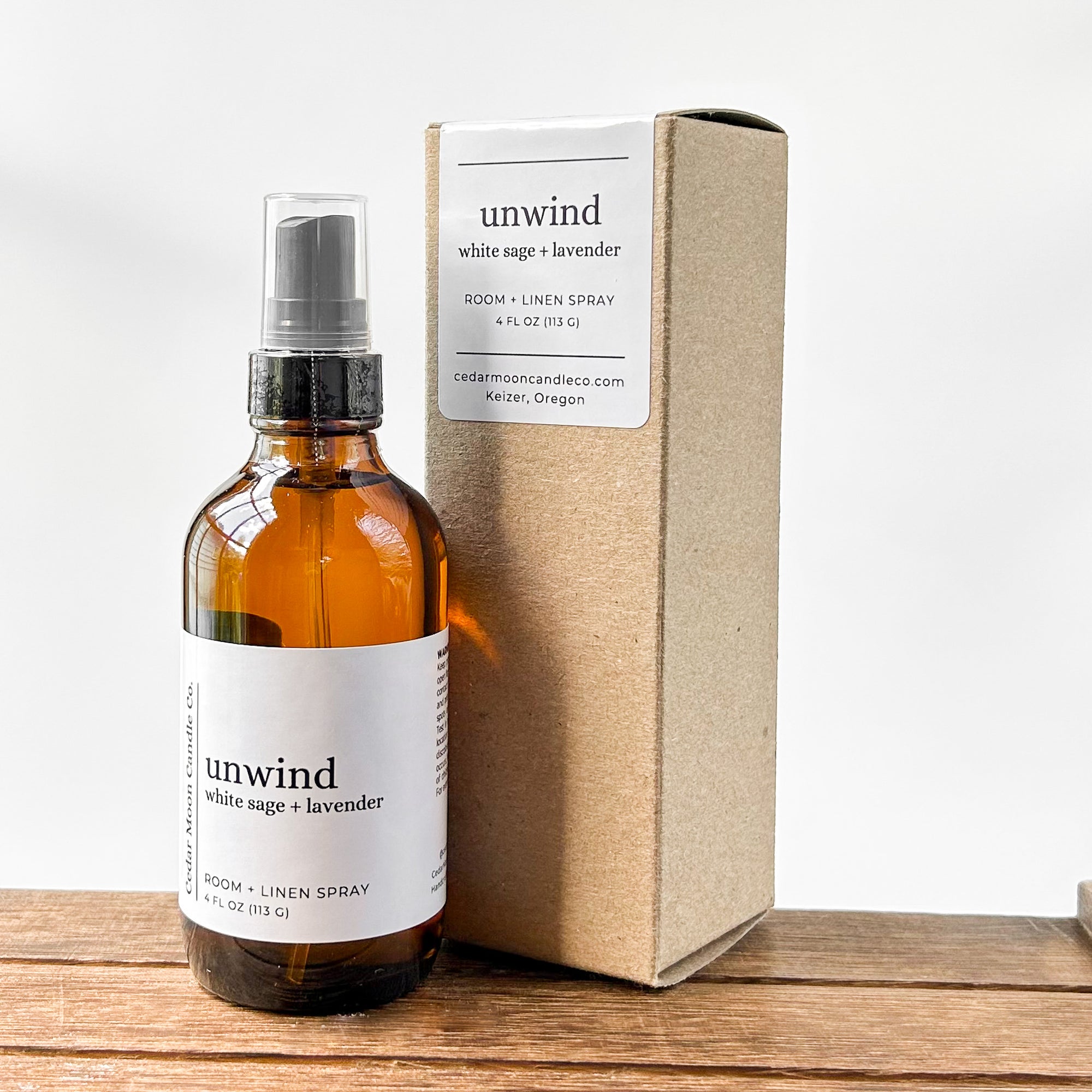 A bottle of room and linen spray labeled 'unwind white sage and lavender' next to a simple cardboard box, resting on a wooden surface with a neutral background.