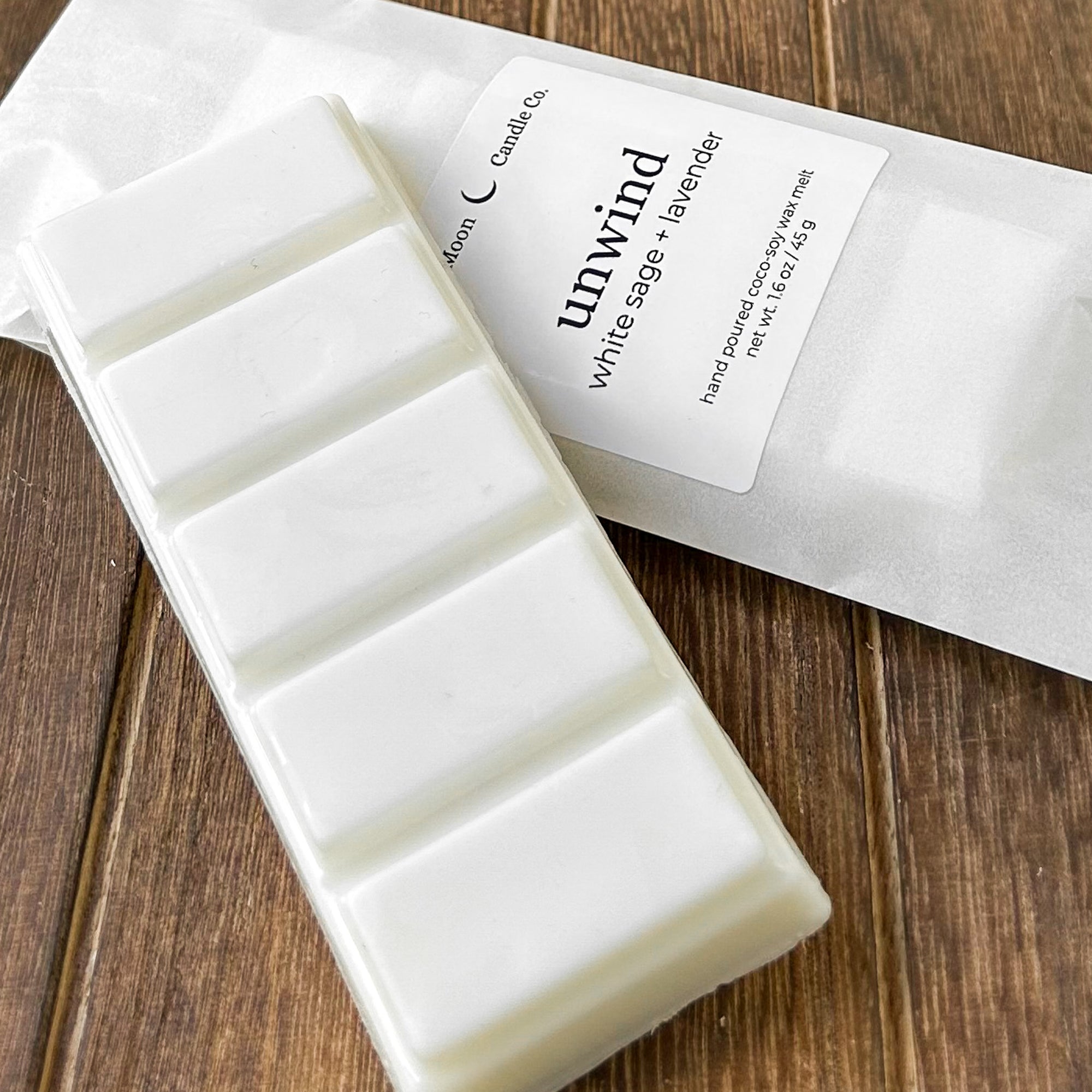 A white wax melt bar labeled 'unwind' with scents of white sage and lavender, resting on a textured wooden surface. The package highlights the product's natural ingredients.