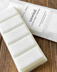 A white wax melt bar labeled 'unwind' with scents of white sage and lavender, resting on a textured wooden surface. The package highlights the product's natural ingredients.