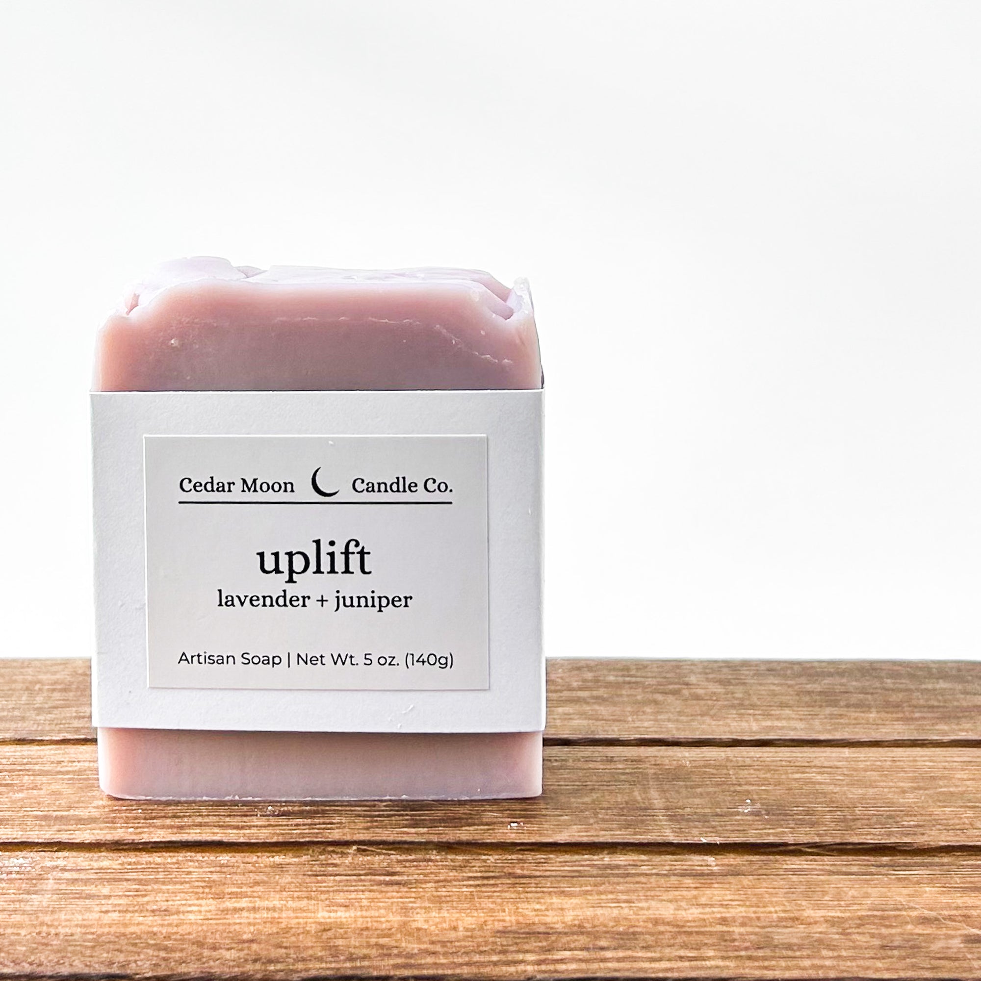 A bar of 'uplift' artisan soap featuring lavender and juniper scents. The soap is pale purple and wrapped in a white paper band placed on a wooden surface against a light background.