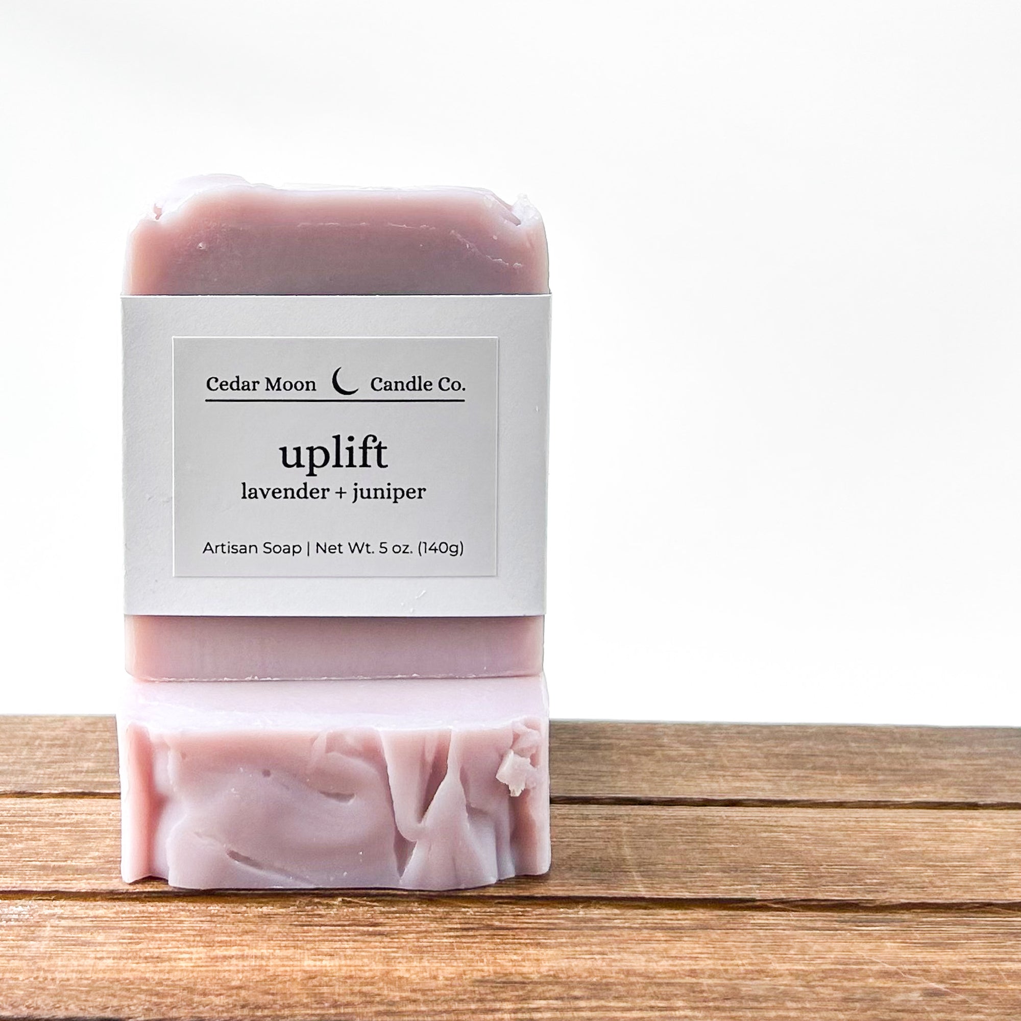A pale purple artisan soap bar labeled 'uplift' featuring lavender and juniper, with a white wrapper displaying the brand 'Cedar Moon Candle Co.' and a wooden surface beneath it.