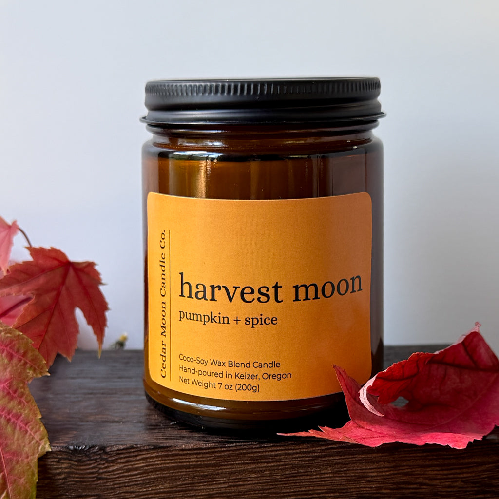 An amber glass jar candle with a label reading 'harvest moon pumpkin + spice' by Cedar Moon Candle Co., styled on a dark wooden surface with red autumn leaves against a light gray background.