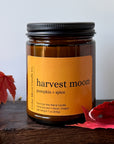 An amber glass jar candle with a label reading 'harvest moon pumpkin + spice' by Cedar Moon Candle Co., styled on a dark wooden surface with red autumn leaves against a light gray background.