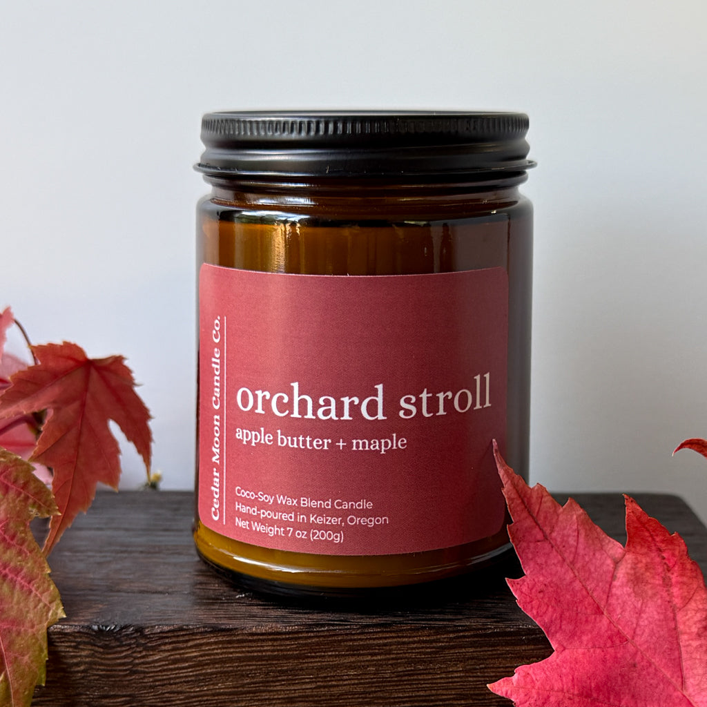 An amber glass jar candle with a black lid reading 'orchard stroll apple butter + maple' by Cedar Moon Candle Co., styled on a dark wooden surface with red autumn leaves against a light gray background.