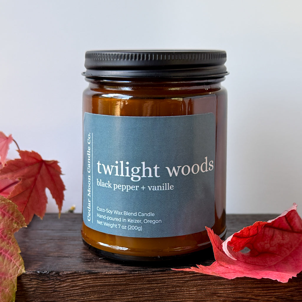 An amber glass jar candle with a reading 'twilight woods black pepper + vanille' by Cedar Moon Candle Co., displayed on a dark wooden surface with red autumn leaves against a light gray background.