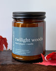 An amber glass jar candle with a reading 'twilight woods black pepper + vanille' by Cedar Moon Candle Co., displayed on a dark wooden surface with red autumn leaves against a light gray background.