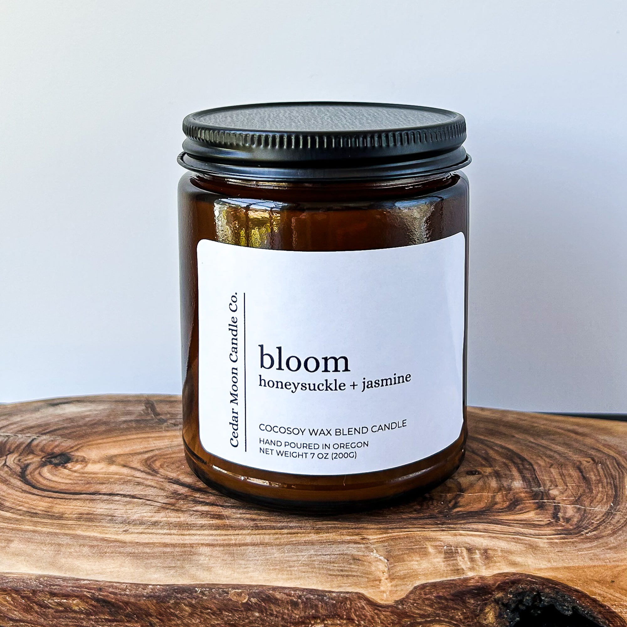 A close-up of a brown glass candle jar labeled 'bloom' with scents of honeysuckle and jasmine, placed on a wooden surface. The candle is a 7 oz cocowax blend, hand poured in Oregon.