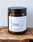 A close-up of a brown glass candle jar labeled 'bloom' with scents of honeysuckle and jasmine, placed on a wooden surface. The candle is a 7 oz cocowax blend, hand poured in Oregon.