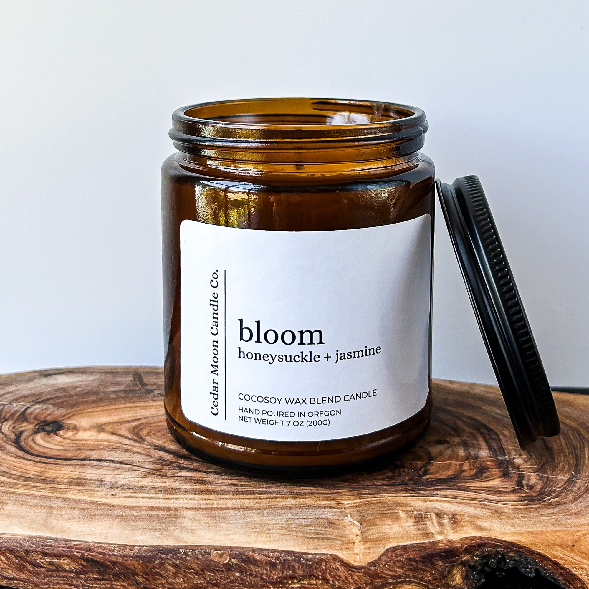 A close-up of a brown glass candle jar labeled 'bloom' with scents of honeysuckle and jasmine, placed on a wooden surface. The candle is a 7 oz cocowax blend, hand poured in Oregon.