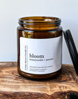 A close-up of a brown glass candle jar labeled 'bloom' with scents of honeysuckle and jasmine, placed on a wooden surface. The candle is a 7 oz cocowax blend, hand poured in Oregon.