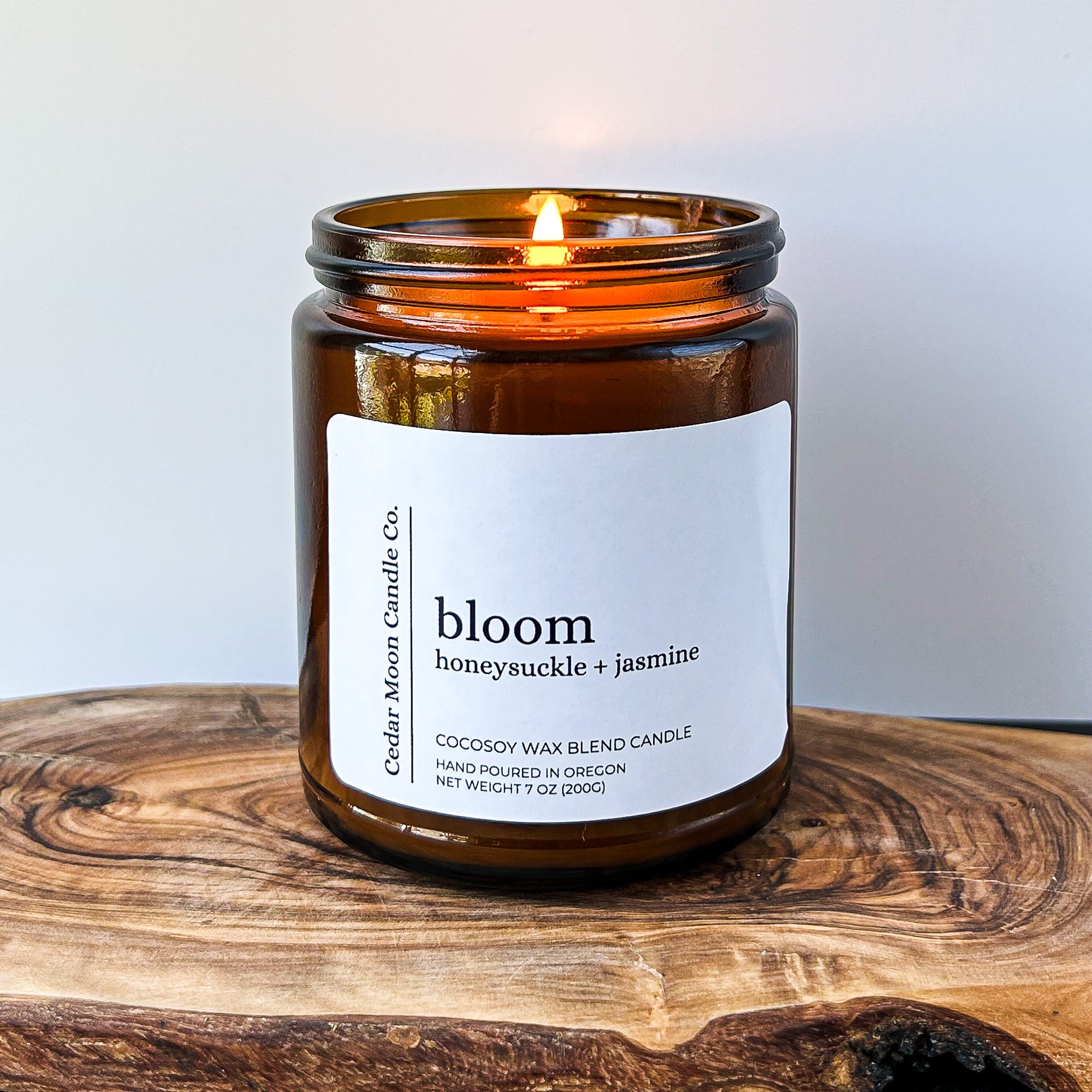 A lit amber jar candle labeled 'bloom honeysuckle  jasmine' sits on a wooden surface. The candle is made from a cocowax blend and was hand poured in Oregon. The flame gently flickers.