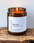 A lit amber jar candle labeled 'bloom honeysuckle  jasmine' sits on a wooden surface. The candle is made from a cocowax blend and was hand poured in Oregon. The flame gently flickers.