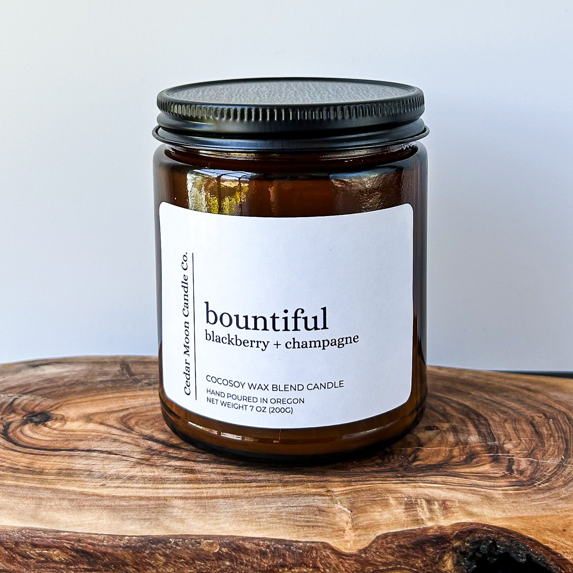 A close-up of a brown glass candle jar labeled 'bountiful' with scents of blackberry and champagne, placed on a wooden surface. The candle is a 7 oz cocowax blend, hand poured in Oregon.