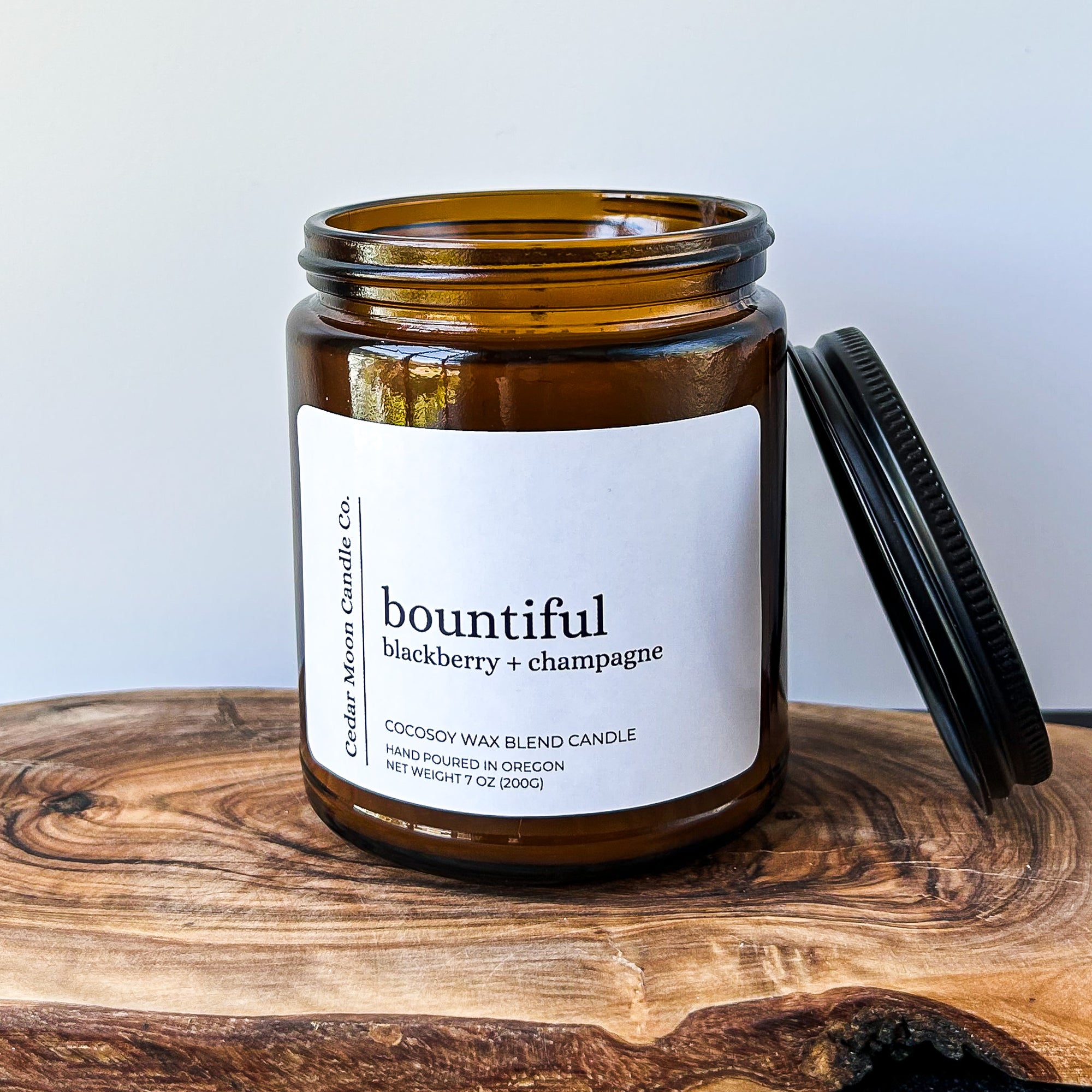 A brown glass candle jar labeled 'bountiful', featuring a blackberry and champagne scent. The jar is sitting on a wooden surface with a black lid positioned next to it.