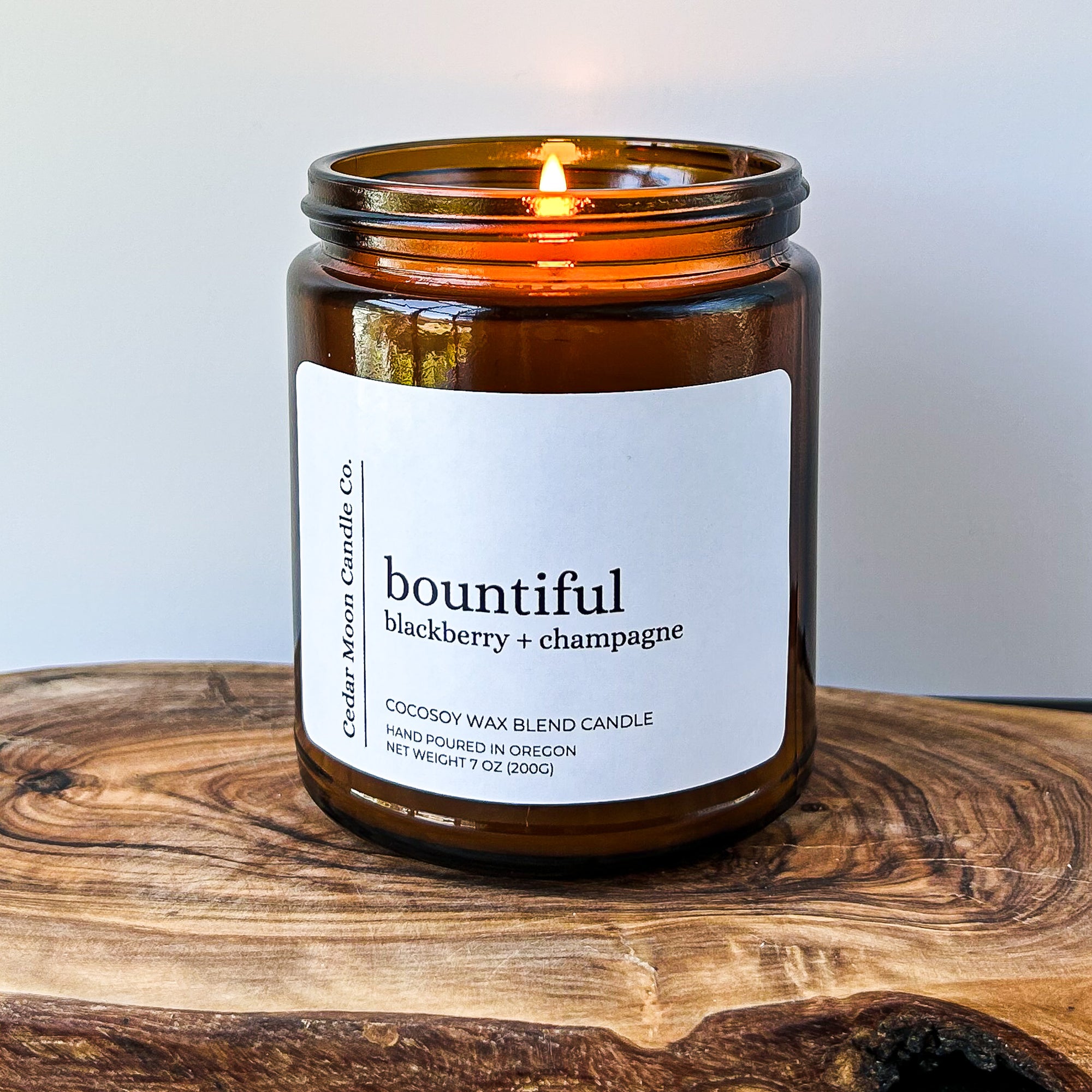 A lit amber jar candle labeled 'bountiful blackberry champagne' sits on a wooden surface. The candle is made from a cocowax blend and was hand poured in Oregon. The flame gently flickers.