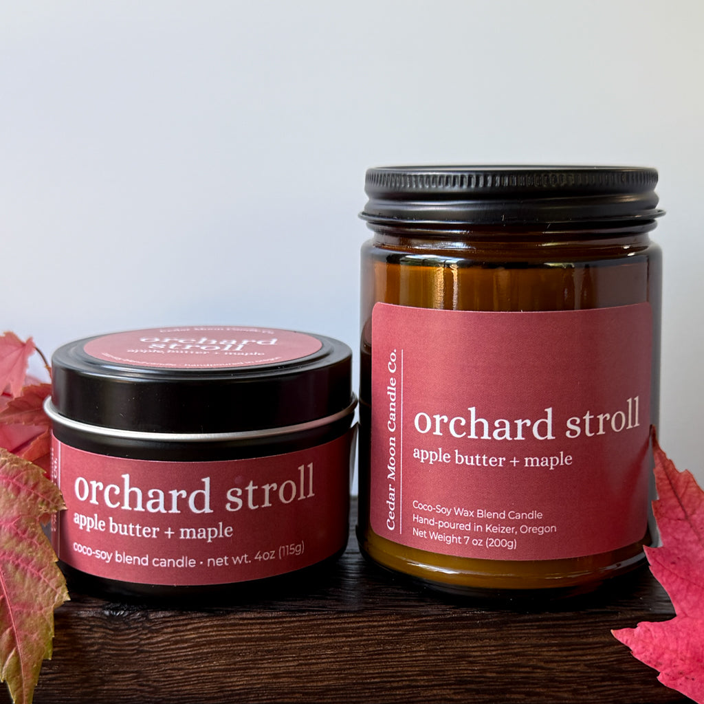Two amber glass and metal candles from Cedar Moon Candle Co. labeled 'orchard stroll apple butter + maple,' displayed on a dark wooden surface with red autumn leaves against a light gray background.