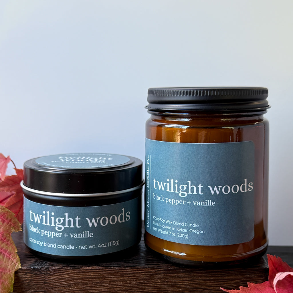 Two Twilight Woods scented candles, one in a round tin and one in a glass jar,  displayed on a dark wooden surface with autumn leaves.