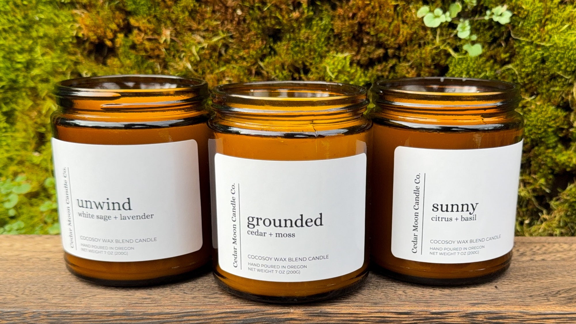 Three candles with labels 'unwind', 'grounded', and 'sunny' on a wooden surface with a green mossy background.