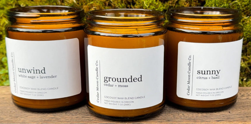 Three candles with labels 'unwind', 'grounded', and 'sunny' on a wooden surface with a green mossy background.