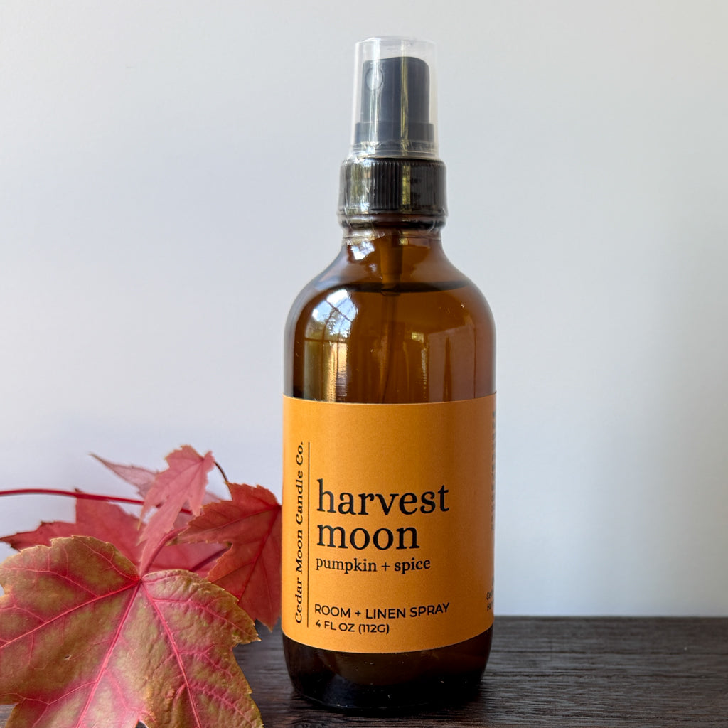 An amber glass room and linen spray bottle with a label reading 'harvest moon pumpkin + spice,' placed on a dark wooden surface with red autumn leaves at the side against a light gray background.