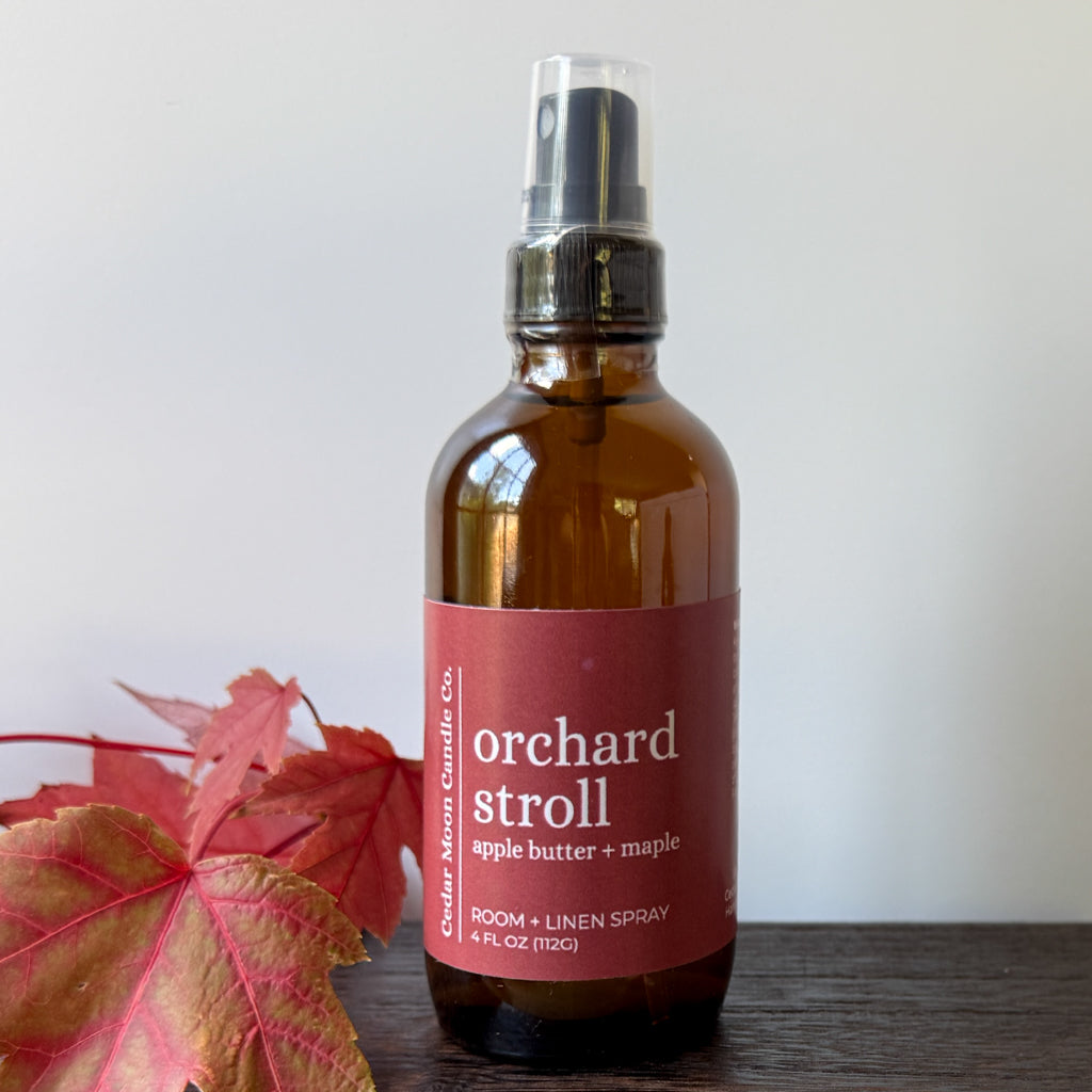 An amber glass room and linen spray bottle with a label reading 'orchard stroll apple butter + maple'. The bottle is placed on a dark wooden surface with red autumn leaves on the left, against a plain off-white wall.