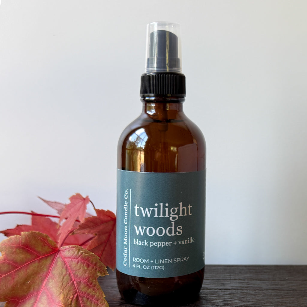 An amber glass room and linen spray bottle with a label reading 'twilight woods black pepper + vanilla' by Cedar Moon Candle Co., set on a dark wooden surface with red autumn leaves against a light gray background.