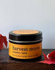A tin candle labeled 'harvest moon pumpkin + spice' sits on a dark wooden surface with red autumn leaves around it against a light gray background.