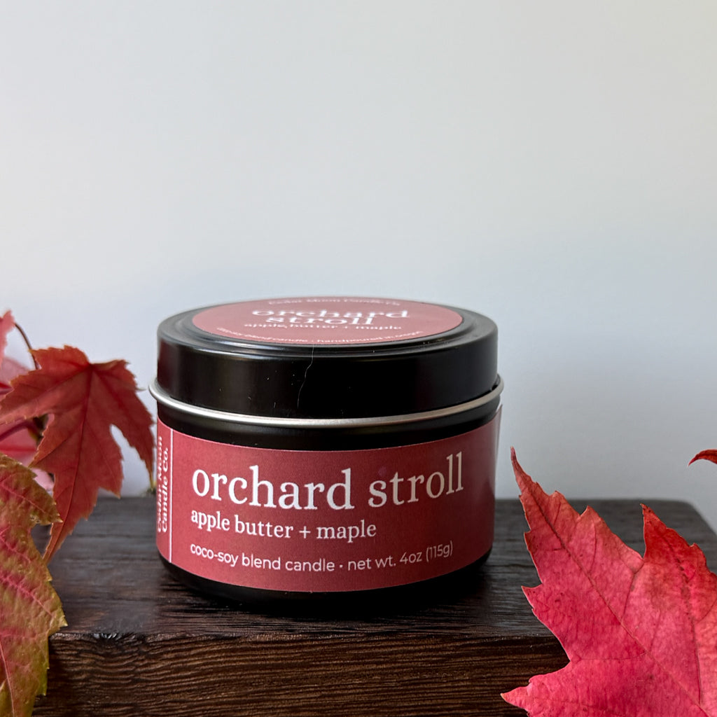 A 6 oz black tin candle labeled 'orchard stroll' by Candle Craft, styled on a dark wooden block with red autumn leaves on either side against a light gray background.