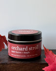 A 6 oz black tin candle labeled 'orchard stroll' by Candle Craft, styled on a dark wooden block with red autumn leaves on either side against a light gray background.