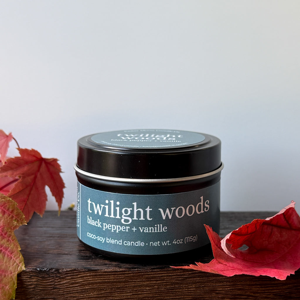 A black candle tin labeled 'twilight woods black pepper + vanille' sits on a dark wooden surface with red autumn leaves around it against a plain light background.