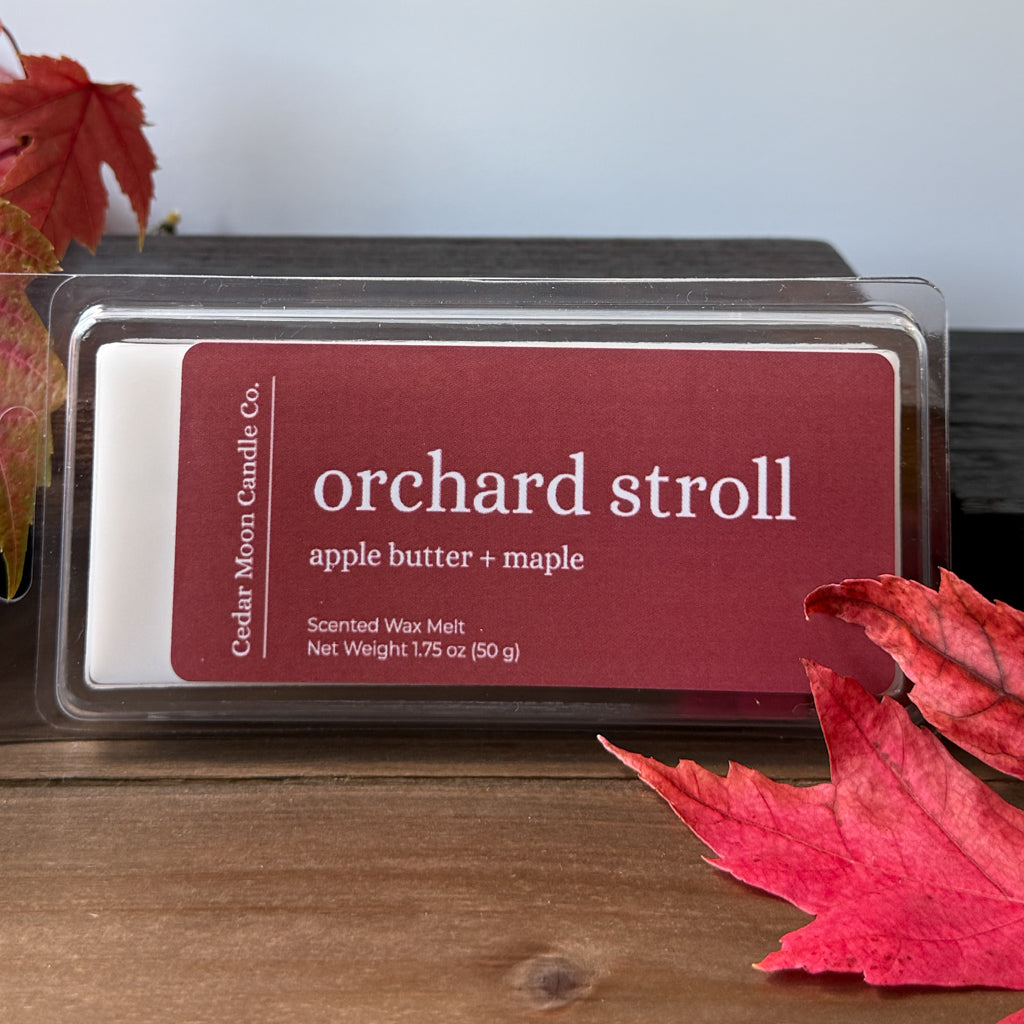 A wax melt package labeled 'orchard stroll' by Cedar Moon Candle Co., placed on a wooden surface with red autumn leaves around it.