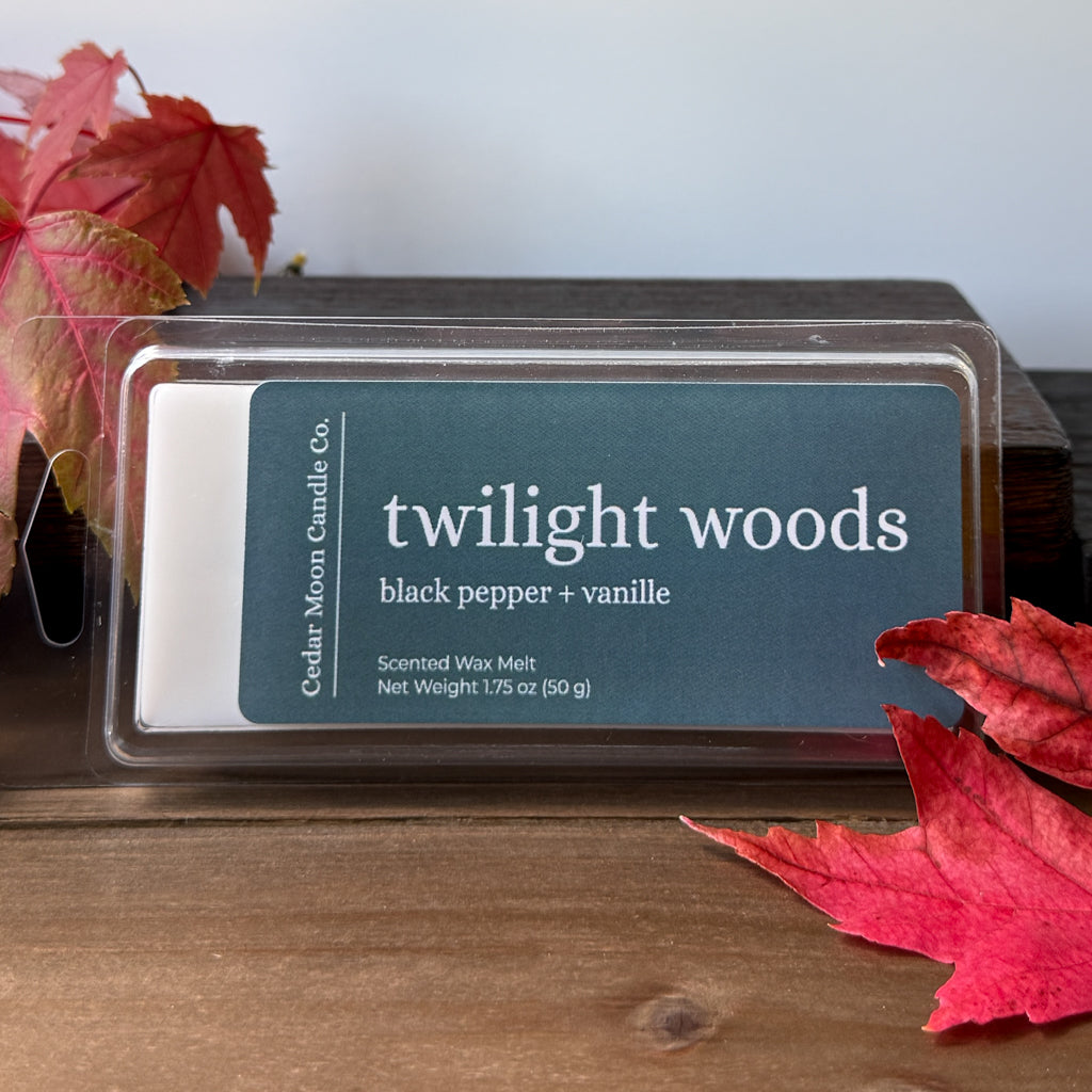 A wax melt package labeled 'twilight woods' by Cedar Moon Candle Co. sits on a wooden surface with red autumn leaves around it.