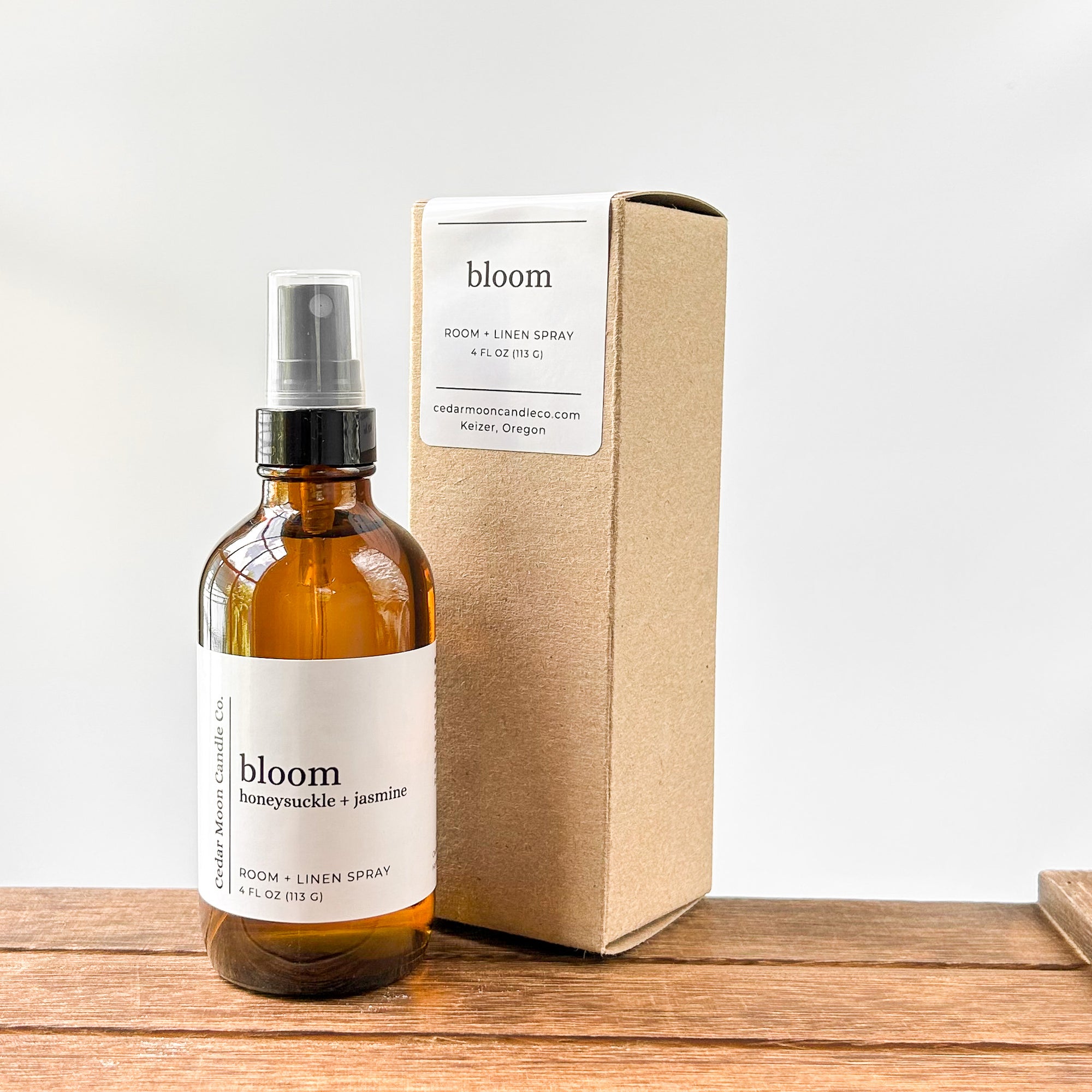 A brown amber glass bottle of 'bloom' room and linen spray with honeysuckle and jasmine scent, placed next to its kraft paper box. The background is a light, neutral color with a wooden surface.