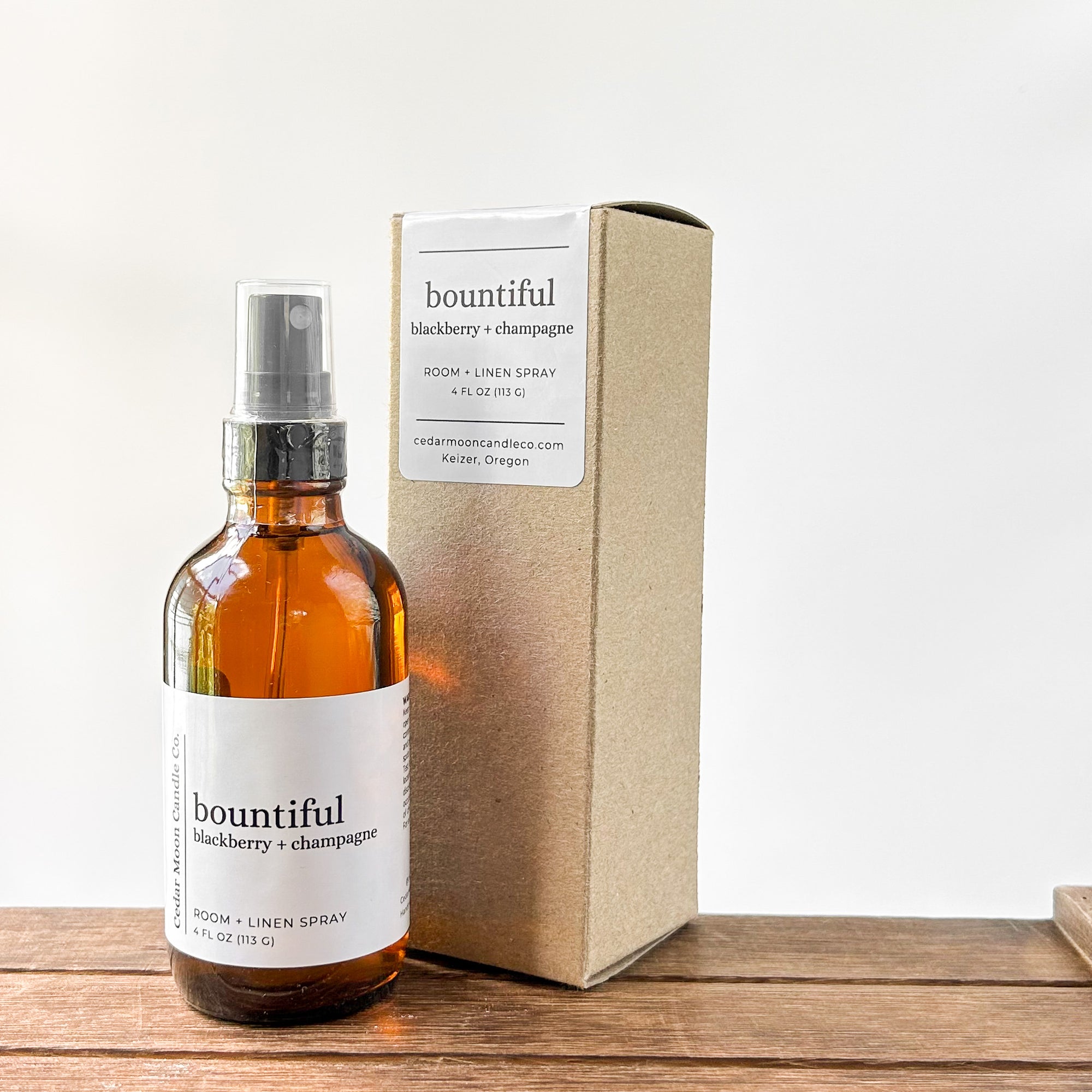 A brown amber glass bottle of 'bountiful' room and linen spray with blackberry and champagne scent, placed next to its kraft paper box. The background is a light, neutral color with a wooden surface.